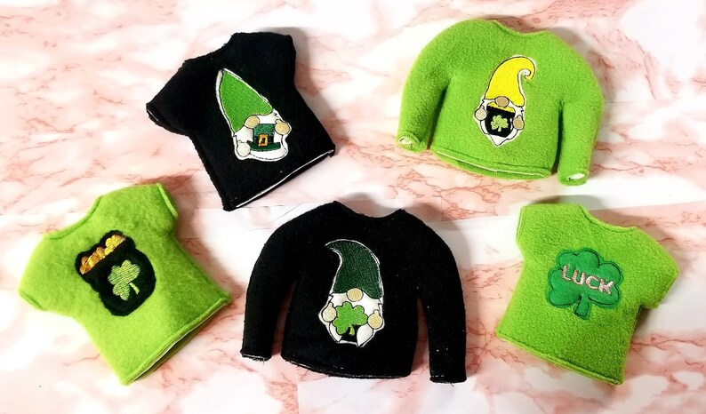 May include: Five felt elf shirts with different St. Patrick's Day designs.  Two shirts are black with a green leprechaun design. Two shirts are green with a gold pot of gold and shamrock design. One shirt is green with the word "LUCK" and a shamrock design.