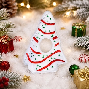 May include: A white, tree-shaped felt ornament featuring a circular cutout in the center. The surface is decorated with embroidered red wavy lines and small green square accents, creating a festive pattern on the front of the decoration.