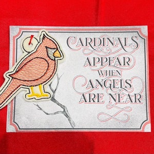 K&ouml;nnte beinhalten: Eine rechteckige Karte mit dem Text Cardinals Appear When Angels Are Near in schwarzer und roter Schrift, begleitet von einem gestickten Kardinalvogel-Ornament mit roten Federn, gelbem Schnabel und gelben F&uuml;&szlig;en an der oberen linken Ecke.