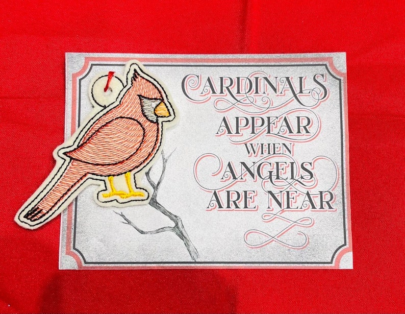 K&ouml;nnte beinhalten: Eine rechteckige Karte mit dem Text Cardinals Appear When Angels Are Near in schwarzer und roter Schrift, begleitet von einem gestickten Kardinalvogel-Ornament mit roten Federn, gelbem Schnabel und gelben F&uuml;&szlig;en an der oberen linken Ecke.