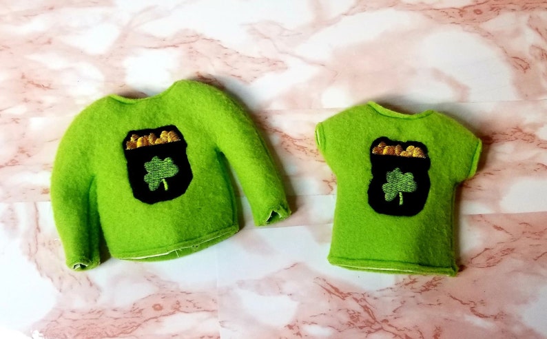 May include: Two green felt shirts with a gold pot of coins and a shamrock design. The larger shirt has long sleeves and the smaller shirt has short sleeves.