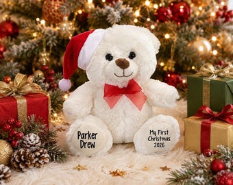 May include: A white plush teddy bear wearing a red and white Santa hat and a red sheer ribbon bow around its neck. The bear has the text Parker Drew on its left foot and My First Christmas 2026 on its right foot in black lettering.