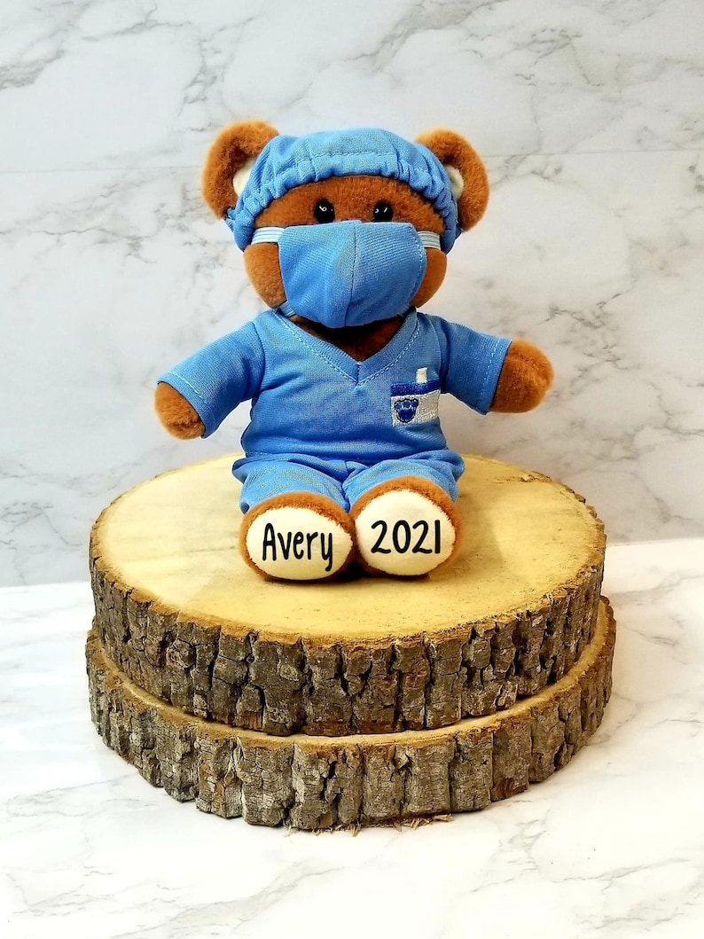 May include: A brown teddy bear wearing a blue surgical scrub top and a blue face mask. The bear has a name tag on its chest and the words "Avery 2021" on its feet. The bear is sitting on a wooden log.
