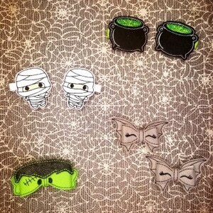 May include: A collection of Halloween-themed hair clips on a spiderweb background. The clips include two mummy figures, two witch's cauldrons, two bat-wing bows, and a Frankenstein's monster bow. The colors are black, white, green, and gray.
