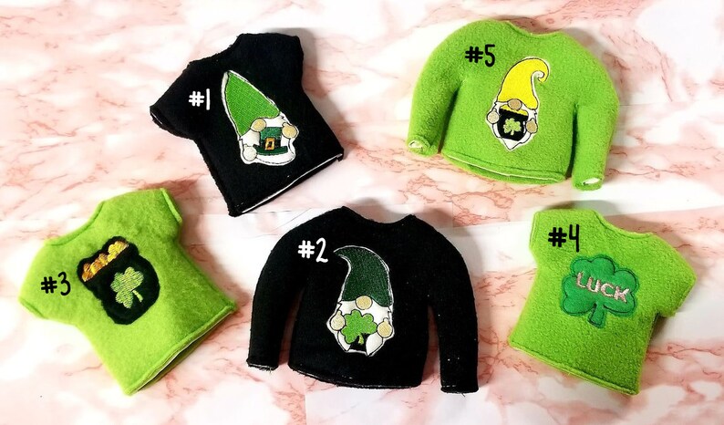 May include: Ten elf-sized St. Patrick's Day shirts and sweaters in green and black with various designs including a gnome, a pot of gold, and a shamrock. The shirts are numbered 1 through 5.