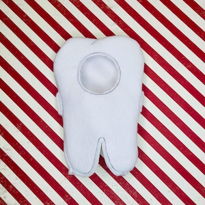 May include: A white plush tooth with a circular detail and black stitching, set against a red and white striped background. The tooth-shaped pillow has a rounded top and two roots.