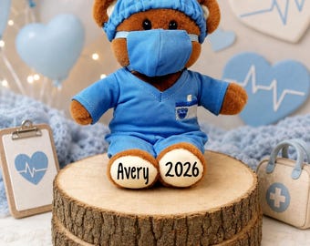 May include: A brown plush teddy bear dressed in a blue medical scrub top, matching scrub cap, and a blue face mask. The bear has the name Avery and the year 2026 printed on its cream-colored foot pads, with a small blue paw print on its chest.