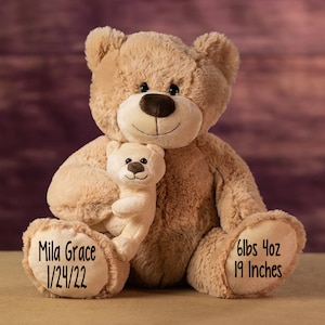May include: A large, plush tan teddy bear holding a smaller, cream-colored teddy bear. The larger bear has the text "Mila Grace 1/24/22" on one paw and "6lbs 4oz 19 Inches" on the other. The bears are set against a blurred, purple and brown background.