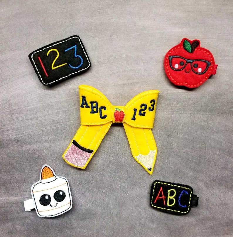 May include: A collection of school-themed felt hair clips. Includes a yellow pencil bow with "ABC 123" text, a red apple with glasses, a glue bottle, and two black chalkboard clips with numbers and letters.