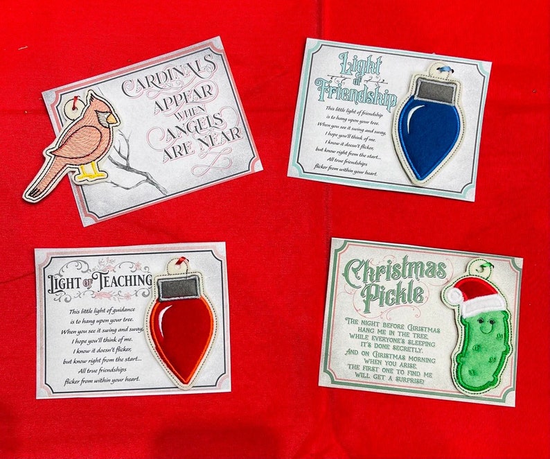 K&ouml;nnte beinhalten: Vier rechteckige Karten mit gestickten Ornamenten: ein roter Kardinal mit dem Text Cardinals Appear When Angels Are Near, eine blaue Gl&uuml;hbirne mit Light of Friendship, eine rote Gl&uuml;hbirne mit Light of Teaching und eine Gew&uuml;rzgurke mit Christmas Pickle.