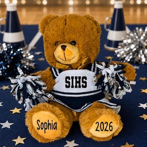 May include: A brown plush teddy bear wearing a navy and white cheerleader uniform with the text SIHS printed on the chest. The bear holds two pom-poms, and its feet are personalized with the name Sophia on one and the year 2026 on the other.