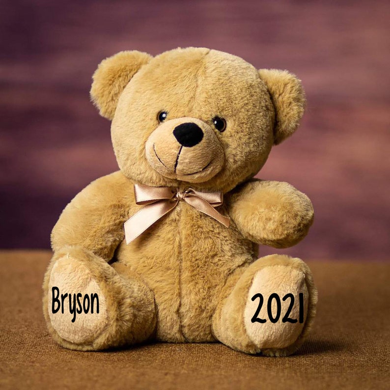 Pu&ograve; includere: Un orsacchiotto di peluche marrone con un fiocco marrone intorno al collo. L'orso &egrave; seduto su una superficie marrone. La zampa sinistra dell'orso ha il nome "Bryson" scritto su di essa e la zampa destra ha l'anno "2021" scritto su di essa.