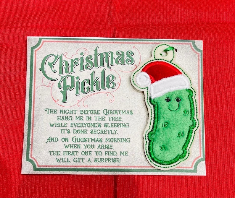 K&ouml;nnte beinhalten: Eine rechteckige Karte mit dem Text Christmas Pickle und einem Gedicht, dazu ein gestickter gr&uuml;ner Gurkenanh&auml;nger mit einer rot-wei&szlig;en Weihnachtsmannm&uuml;tze. Die Karte hat einen dekorativen Rand und liegt auf einem einfarbig roten Hintergrund.