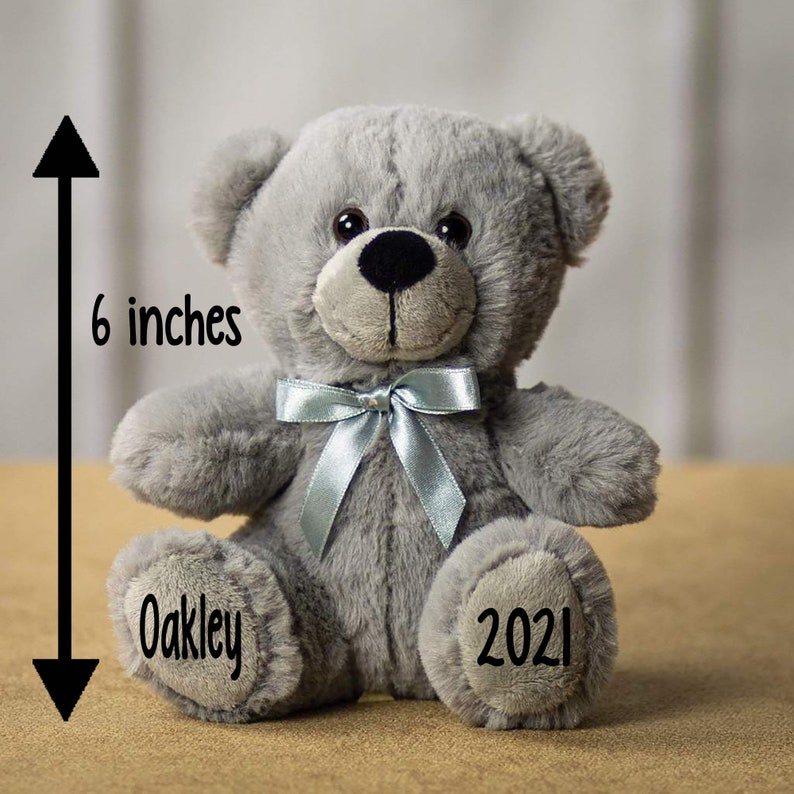 Pu&ograve; includere: Un orsacchiotto di peluche grigio con un nastro blu intorno al collo. L'orso &egrave; alto 15 cm e ha il nome "Oakley" e l'anno "2021" ricamato sulle zampe.