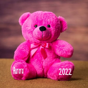 Pu&ograve; includere: Un orsacchiotto rosa con un fiocco rosa, seduto su una superficie marrone. L'orsacchiotto ha il nome "Aurora" ricamato su una zampa e l'anno "2022" sull'altra.