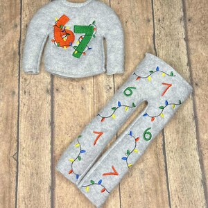 May include: A gray felt sweater and pants set with the numbers "67" embroidered on the sweater and the numbers "6" and "7" on the pants. The numbers are decorated with colorful string lights.