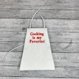 May include: White apron with red embroidered text that reads "Cooking is my Favorite!" The apron has a simple design with a neck loop and waist ties. The apron is made of a soft, felt-like material.