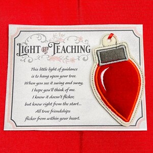 K&ouml;nnte beinhalten: Ein Weihnachtsbaumschmuck aus Filz in Form einer roten Gl&uuml;hbirne mit grauem Oberteil und roter Bandschlaufe, befestigt auf einer rechteckigen Karte mit dem Text LIGHT OF TEACHING und einem Gedicht &uuml;ber Freundschaft, umrahmt von einer floralen Bord&uuml;re.