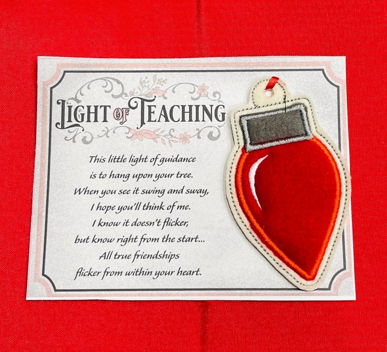 K&ouml;nnte beinhalten: Ein Weihnachtsbaumschmuck aus Filz in Form einer roten Gl&uuml;hbirne mit grauem Oberteil und roter Bandschlaufe, befestigt auf einer rechteckigen Karte mit dem Text LIGHT OF TEACHING und einem Gedicht &uuml;ber Freundschaft, umrahmt von einer floralen Bord&uuml;re.