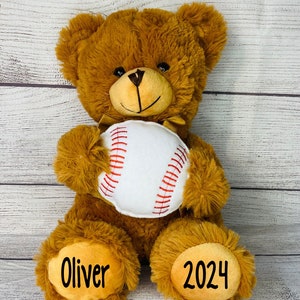 May include: A brown teddy bear holding a white baseball with red stitching. The bear's paws have the names "Oliver" and "2024" written on them.