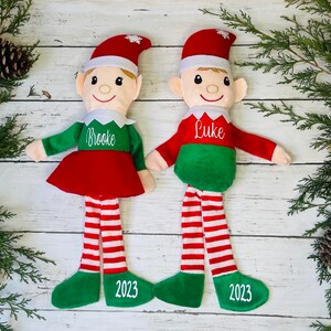 May include: Two personalized Christmas elves with red and white striped legs and green felt hats. The elves are wearing green shirts with the names "Brooke" and "Luke" embroidered on them. The elves are standing on a white wooden surface with the year "2023" embroidered on their feet.