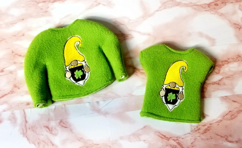 May include: Two green fleece shirts with a gnome holding a pot of gold and a shamrock embroidered on the front. The shirt on the left has long sleeves and the shirt on the right has short sleeves.
