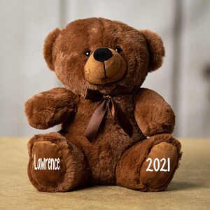 Pu&ograve; includere: Un orsacchiotto di peluche marrone con un nastro marrone intorno al collo. L'orsacchiotto ha il nome "Lawrence" ricamato su un piede e l'anno "2021" ricamato sull'altro piede.