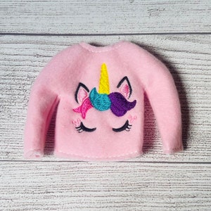 May include: Pink fleece elf-sized sweater with an embroidered unicorn design featuring a yellow horn, teal and purple mane, and pink star.