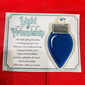 K&ouml;nnte beinhalten: Ein blauer Filz-Weihnachtsbaumschmuck in Gl&uuml;hbirnenform mit grauer Kappe und blauer Bandschlaufe, befestigt auf einer Karte mit dem Text Light of Friendship und einem Gedicht &uuml;ber Freundschaft. Die Karte hat einen dekorativen Rand auf rotem Grund.