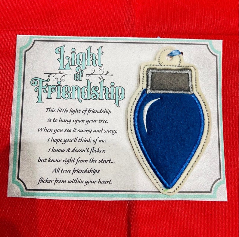 K&ouml;nnte beinhalten: Ein blauer Filz-Weihnachtsbaumschmuck in Gl&uuml;hbirnenform mit grauer Kappe und blauer Bandschlaufe, befestigt auf einer Karte mit dem Text Light of Friendship und einem Gedicht &uuml;ber Freundschaft. Die Karte hat einen dekorativen Rand auf rotem Grund.