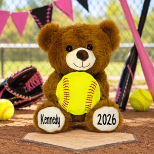 May include: A brown plush teddy bear holding a bright yellow softball with red stitching. The bear has white paw pads with the name Kennedy printed on the left foot and the year 2026 printed on the right foot in a black sans-serif font.