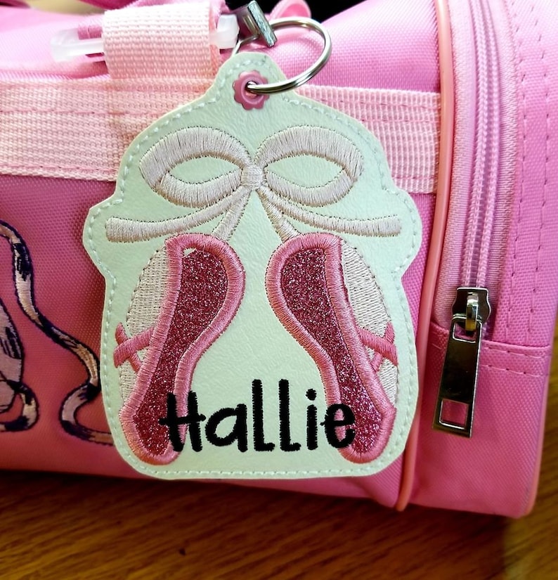 May include: A light green fabric tag with pink ballet shoes embroidered on it. The tag has a white ribbon and the name "Hallie" embroidered in black.