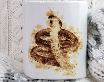 May include: White ceramic mug featuring a watercolor-style illustration of a cobra in shades of brown. The snake is coiled and upright, with a detailed depiction of its scales. The mug has the words "BEANY BEANY" in a circular design.