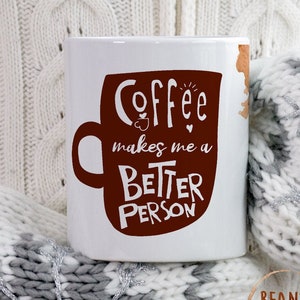 Può includere: Tazza da caffè in ceramica bianca con un design a tazza di caffè marrone e la scritta "Coffee makes me a BETTER PERSON" in bianco. La tazza è appoggiata su una calda coperta di maglia bianca e grigia.