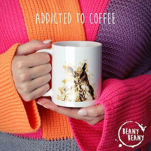May include: White ceramic coffee mug with a watercolor owl design and the text "ADDICTED TO COFFEE". The mug also has the "BEANY BEANY" logo. The mug is held by a person wearing a colorful sweater.