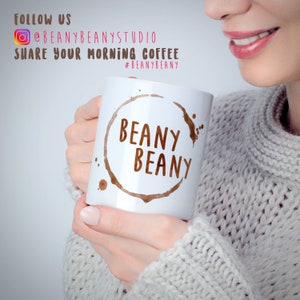 May include: White ceramic coffee mug with the words "BEANY BEANY" in brown, surrounded by a coffee stain design. The mug is held by a person wearing a gray sweater. Text in the background reads "FOLLOW US @BEANYBEANYSTUDIO SHARE YOUR MORNING COFFEE #BEANYBEANY".