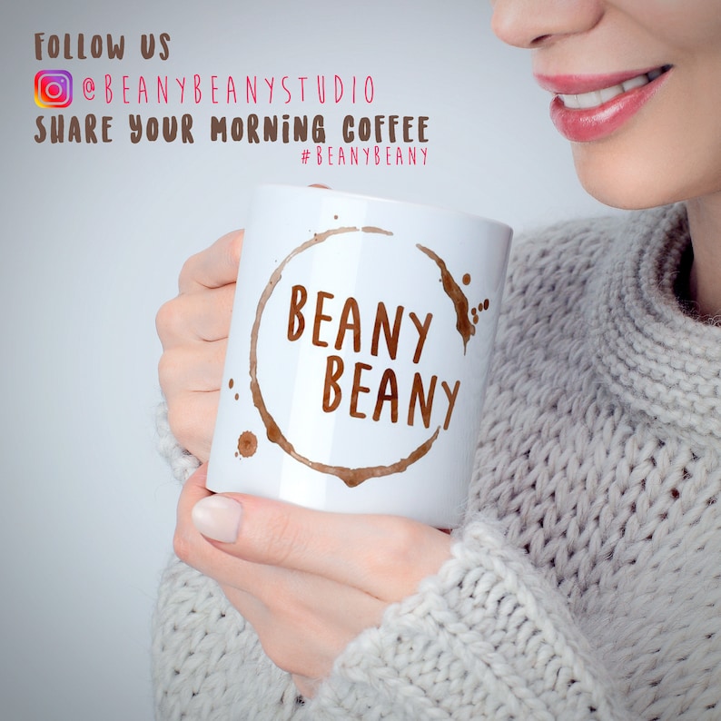 May include: White ceramic coffee mug with the words "BEANY BEANY" in brown, surrounded by a coffee stain design. The mug is held by a person wearing a gray sweater. Text in the background reads "FOLLOW US @BEANYBEANYSTUDIO SHARE YOUR MORNING COFFEE #BEANYBEANY".