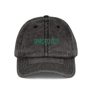 May include: A dark gray baseball cap with the words "SPACED OUT" embroidered in green across the front. The cap has a curved brim and an adjustable strap at the back.