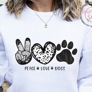 May include: White sweatshirt featuring a black and white graphic with the words "Peace Love Dogs". The design includes a peace sign, a heart, and a dog paw print. The sweatshirt has a crew neck.