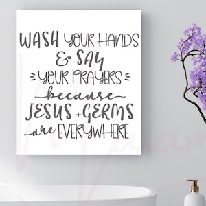 May include: A white square wall sign featuring dark grey text that reads: WASH YOUR HANDS & SAY YOUR PRAYERS because JESUS + GERMS are EVERYWHERE. The sign is displayed in a bathroom setting with a white bathtub and a tree branch with purple flowers.