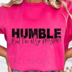 以下が含まれることがあります： 黒色の「HUMBLE But I'm also pressure」の文字が入った鮮やかなピンク色のスウェットシャツ。スウェットシャツは無地で長袖です。「DIGITAL DESIGNS」の文字が下部に印刷されています。