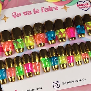 Peut inclure: Ensemble de faux ongles avec des pointes dorées et des couleurs néon vives, dont vert, rose, bleu et jaune. Les ongles sont disposés sur une carte avec le texte "Ça va le faire" et des identifiants de réseaux sociaux.