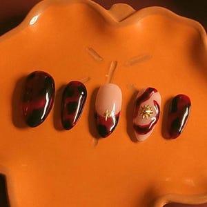 Peut inclure: Un plat orange en forme de feuille présente cinq faux ongles. Les ongles présentent divers motifs, notamment un motif écailles de tortue rouge foncé, une base nude avec un bout français rouge et des accents dorés. Le fond est brun rougeâtre foncé.