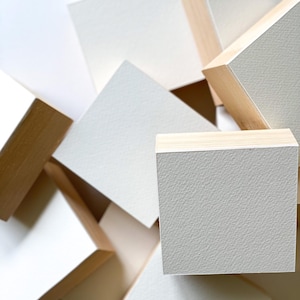 Könnte beinhalten: Eine abstrakte Komposition aus Holzblöcken mit strukturierten, cremefarbenen Oberflächen. Die Blöcke sind in einem scheinbar zufälligen Muster angeordnet, wobei einige die natürliche Holzmaserung an ihren Seiten zeigen. Das Bild hat eine neutrale Farbpalette und einen Fokus auf Textur und Form.