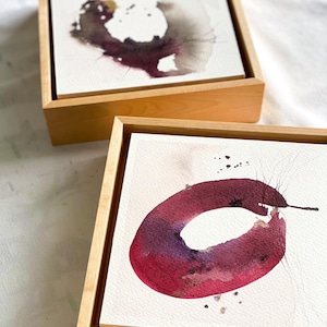May include: Two abstract watercolor paintings in wooden frames. One features a dark, organic shape with gold accents. The other displays a deep red, circular form with a lighter center and delicate lines.
