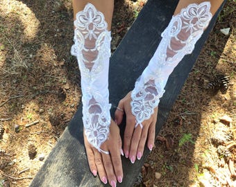 Puede incluir: Un par de guantes de encaje blancos de longitud codo con bordados florales y detalles de perlas. Los guantes tienen un diseño sin dedos con un pequeño lazo de cinta en la muñeca.