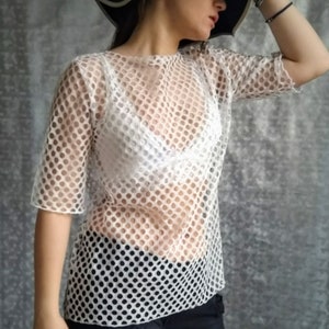 May include: A white mesh top with a round neckline and short sleeves. The top is sheer and has a small, repeating pattern of circles. The model is wearing the top over a white bra.