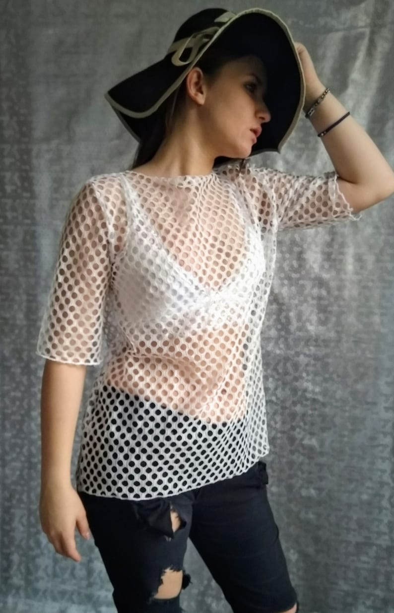 May include: A white mesh top with a round neckline and short sleeves. The top is sheer and has a small, repeating pattern of circles. The model is wearing the top over a white bra.