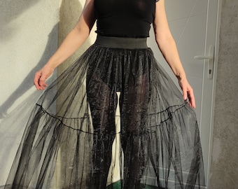 May include: A woman wearing a black top and a long black tulle skirt with multiple layers. The skirt is sheer and flows around her legs. She is standing in front of a white door.
