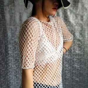 May include: A white mesh top with a round pattern. The top is sheer and has short sleeves. The model is wearing a white bra underneath the top.